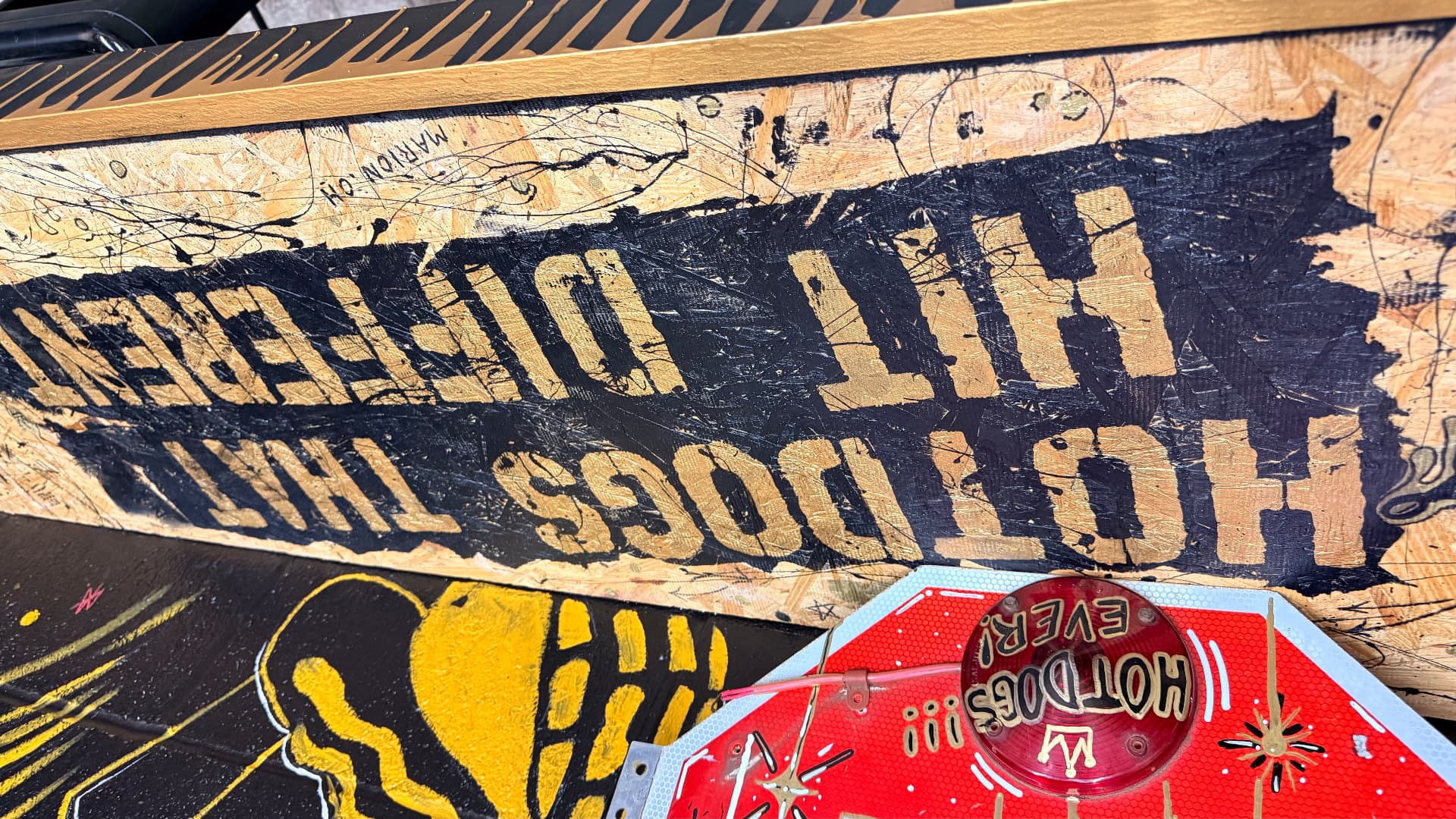 Gold stencil sign reading Hot Dogs That Hit Different, Marion OH, on raw OSB board